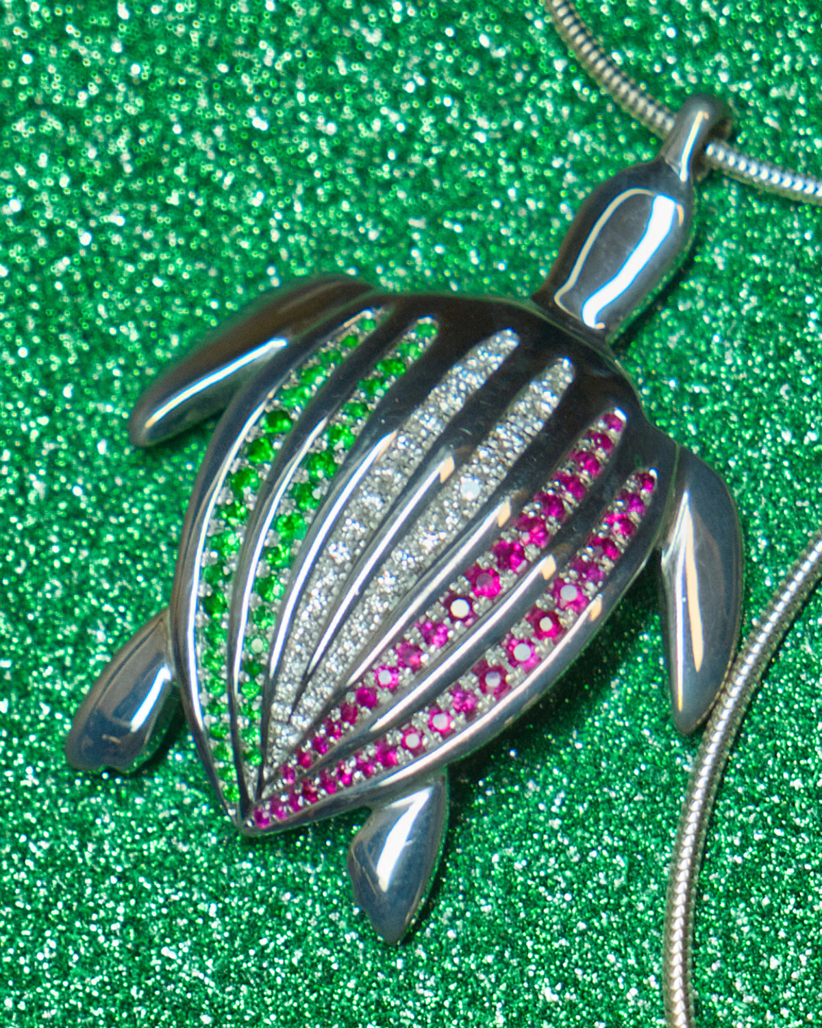 Le pendentif Totem Luth Italia, tsavorites, diamants et rubis sur argent massif, en gros plan sur un fond vert pailleté | Honu
