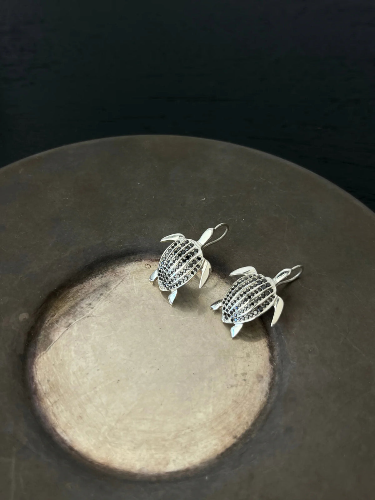 Les boucles d'oreilles Totem Luth, diamants noirs sur argent massif, posées dans une coupelle en métal patiné | Honu