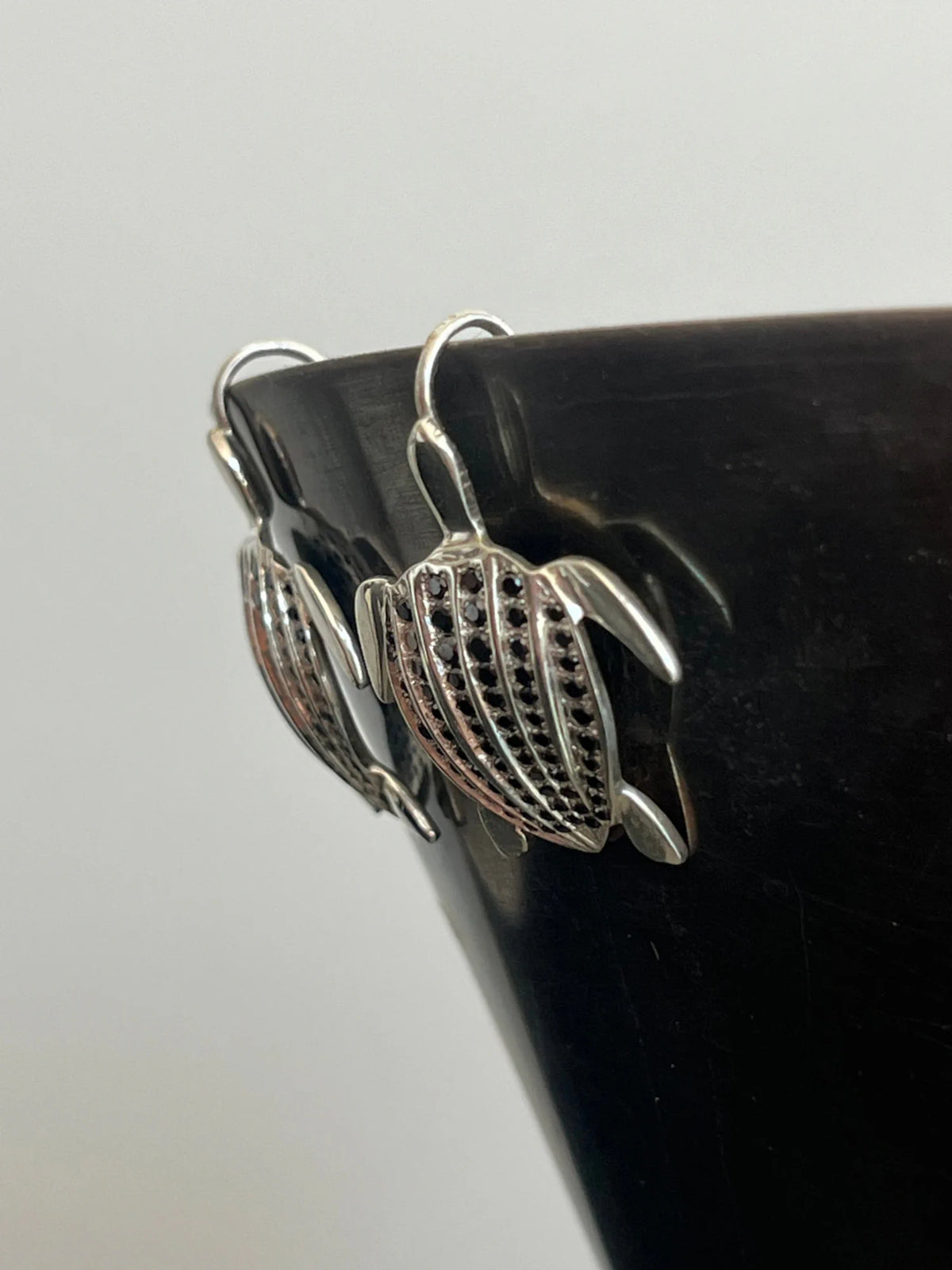 Les boucles d'oreilles Totem Luth, diamants noirs sur argent massif, en gros plan accrochées au bord d'un bol noir | Honu