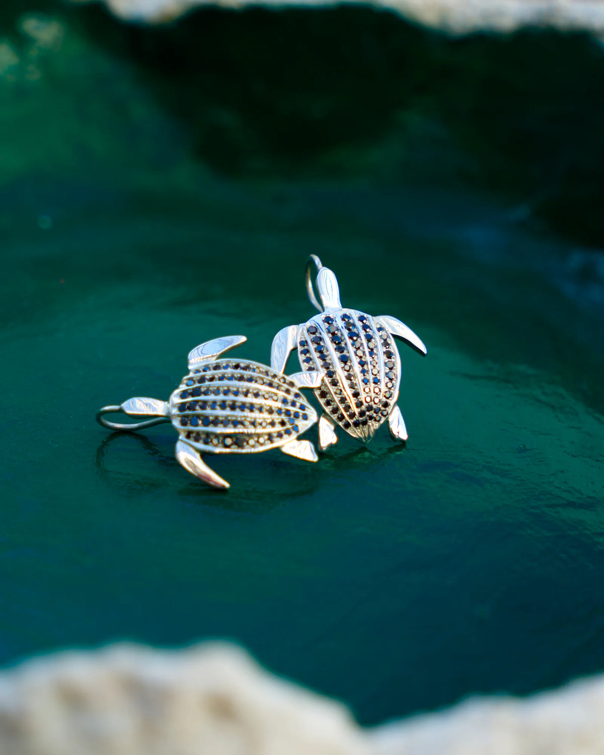 Les boucles d'oreilles Totem Luth, diamants noirs sur argent massif, posées au fond d'une coupelle vert émeraude, corail blanc en premier plan | Honu