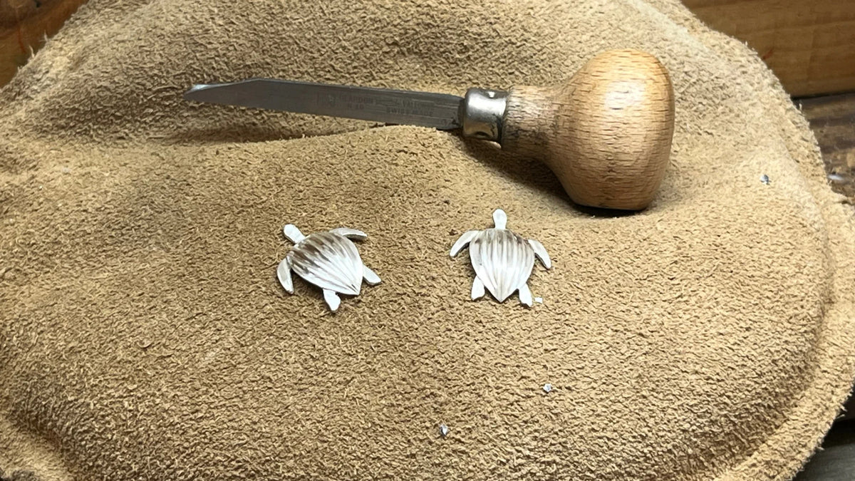 Gros plan des deux tortues Luth en argent, carènes sculptées une à une, posées sur le coussin de sable près de l'échoppe.