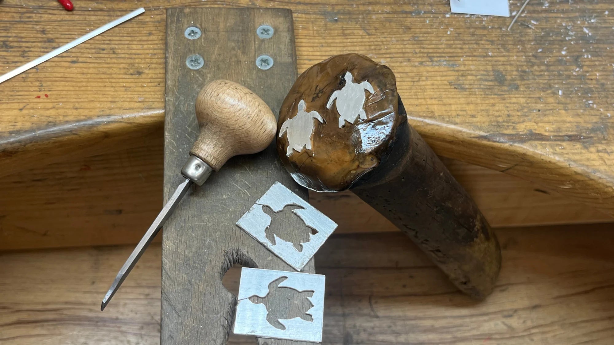 Plan de travail de l'atelier : trois motifs de tortue Luth en argent pris dans la poix, une échoppe et deux plaques d'argent ajourées, près de la cheville en bois.