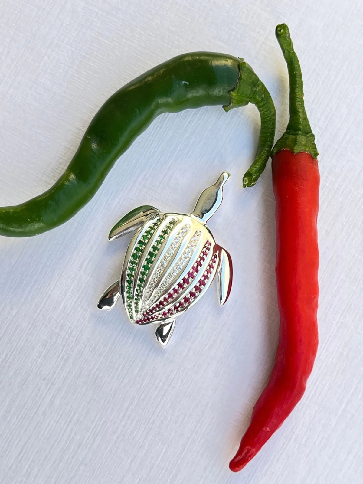 Le pendentif Totem Luth Italia, tsavorites, diamants et rubis sur argent massif, posé entre un piment vert et un piment rouge | Honu