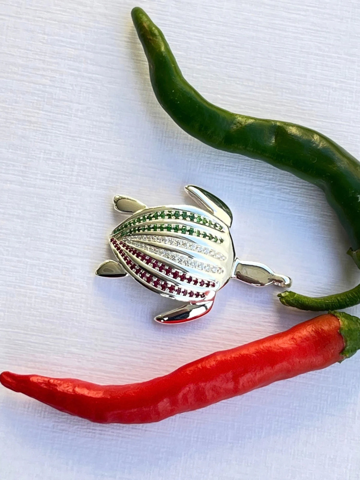 Le pendentif Totem Luth Italia, tsavorites, diamants et rubis sur argent massif, posé à l'horizontale entre piments vert et rouge | Honu
