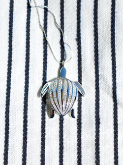 The Totem Luth pendant, resting on a striped piece of fabric with its chain unfurled, from the Totem Luth | Honu Collection